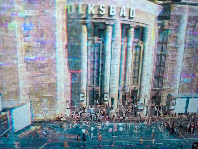 Eine große historische Gebäudefassade mit der Aufschrift „VOLKSBAD“ und hohen Säulen. Vor dem Eingang befindet sich ein temporäres Wasserbecken, in dem zahlreiche Menschen baden, sitzen und sich aufhalten.